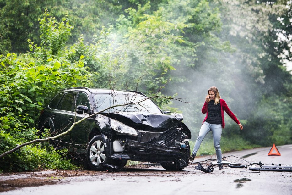 An angry young woman with smartphone by the damaged car after a car accident.